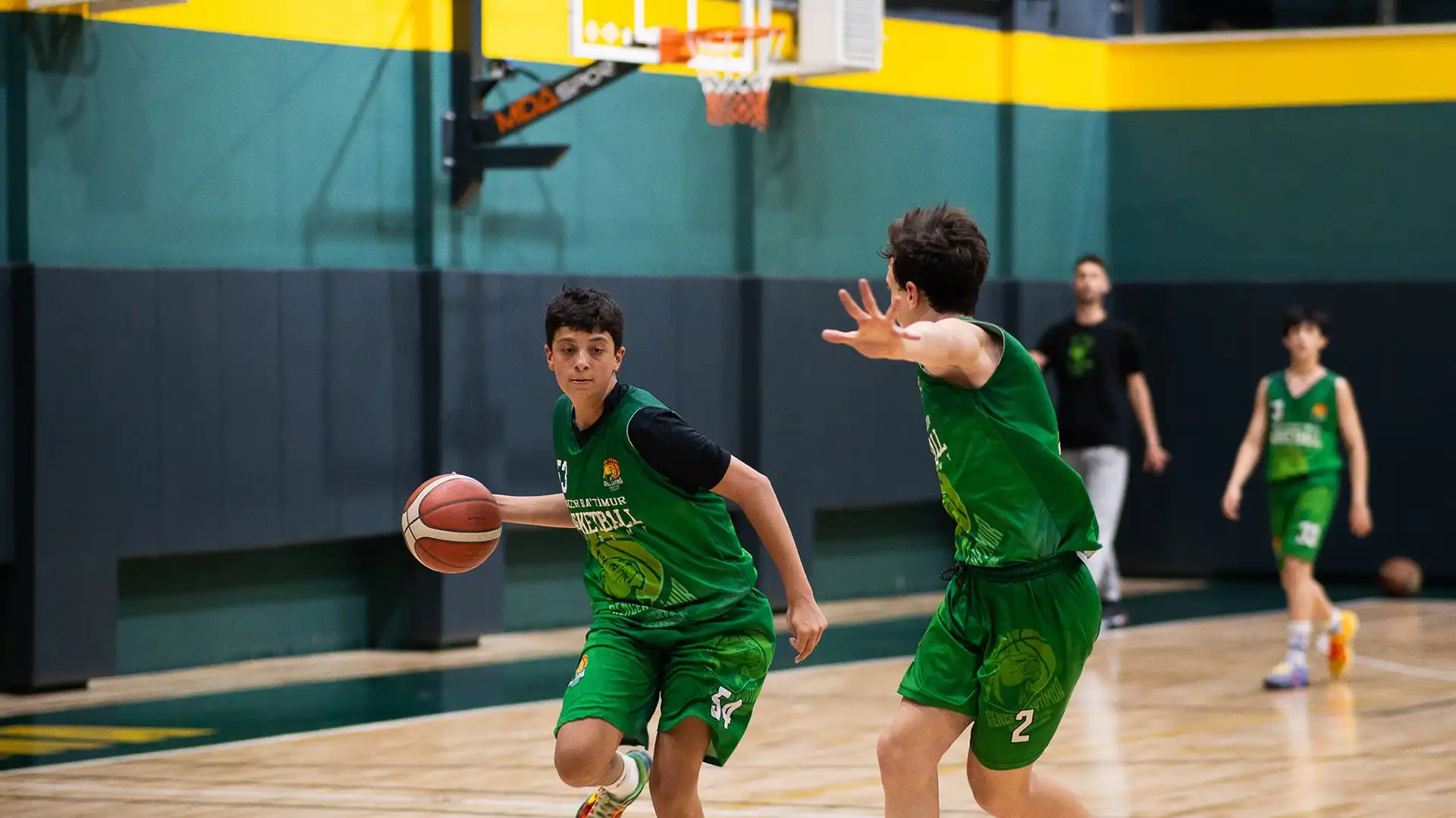 Basketbol oynayan genç sporcular sahada top sürerken ve savunma yaparken, Gencer Baytimur Basketbol Akademisi